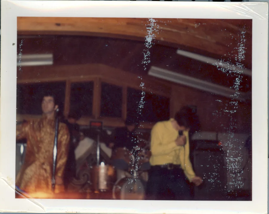 Color print with heavy silver emulsion damage: on stage under a wood-paneled ceiling, a singer in a gold brocade jacket at the microphone at left while another performer in yellow dances at right, drums behind