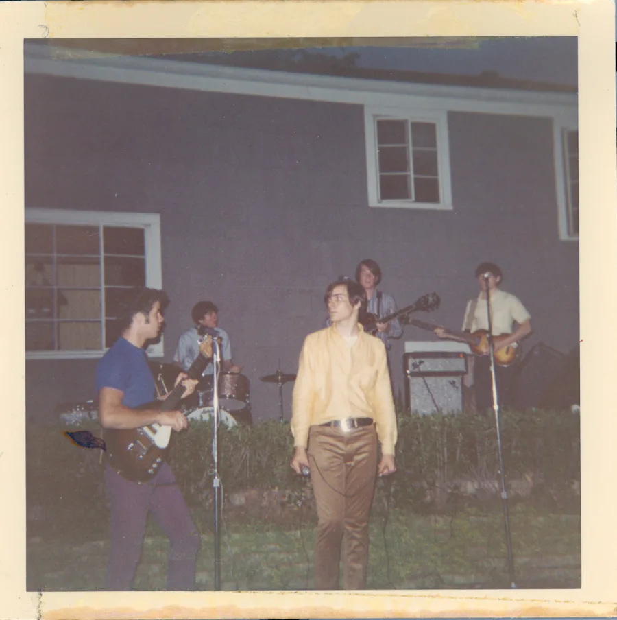Square color print of a backyard performance at dusk: five members playing in front of a gray clapboard house, a guitarist in a blue tee in the foreground, singer in a yellow shirt at center, drummer and two more musicians behind, amps along the hedge