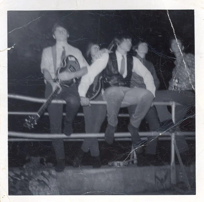 Black-and-white photograph of five boys in ties sitting in a row on a metal railing at night, one cradling a guitar, the print heavily creased and cracked