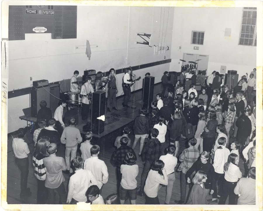 Black-and-white photograph from a balcony of a packed high-school gymnasium dance, HOME and VISITOR scoreboard on the back wall, the band performing on a low stage at left over a dense crowd of teenagers in 1960s dress