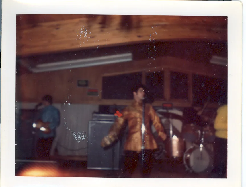 Color print of the singer in the gold brocade jacket mid-move at center stage, arm extended, drum kit at right and a second vocalist at left under the same wood-paneled ceiling
