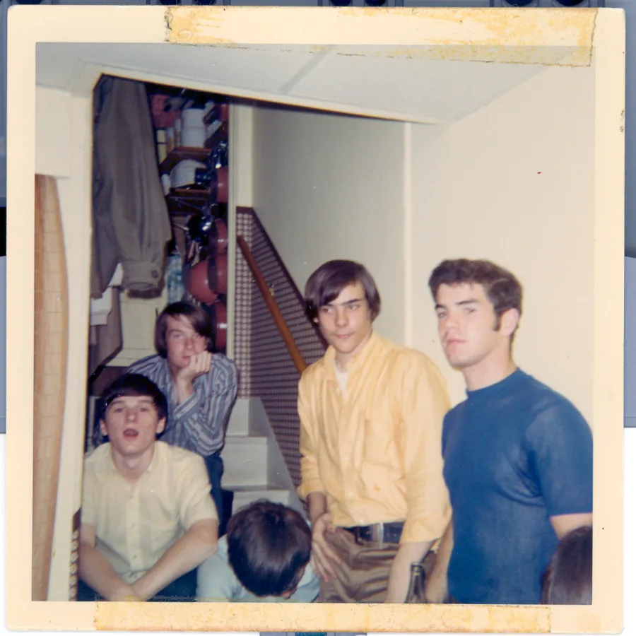 Square color print of five band members crowded on a basement stair landing between sets, in yellow and blue shirts, coats and cluttered shelves visible up the staircase behind them