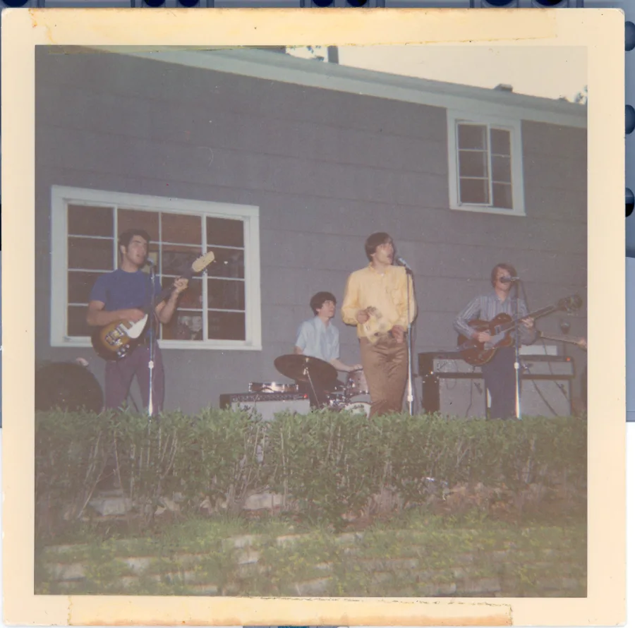 Square color print of the same backyard show from further back: the band on a terrace above a stone retaining wall and hedge, guitarist at left, drummer center, singer in yellow at the microphone, second guitarist at right before the gray house