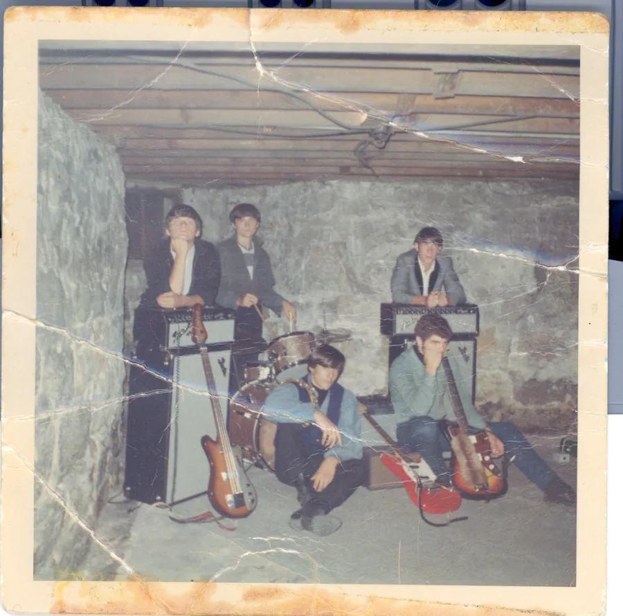 Square color print, heavily creased, of the five posed in a stone-walled basement with Fender amplifiers, a drum kit, and two bass guitars laid across the floor in front