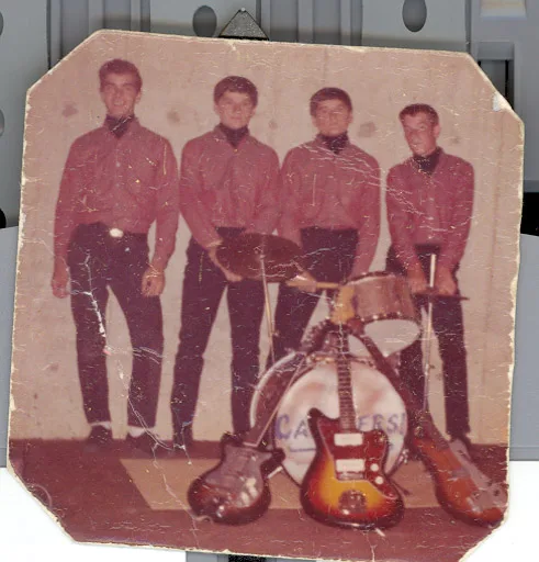 Small color print, corners clipped: four young men in matching dark red shirts and black slacks behind a drum kit, three electric guitars propped against it, the drum head hand-lettered with an earlier band name now worn illegible